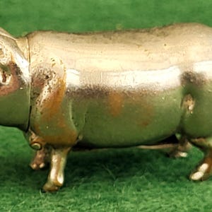 Puede incluir: Pequeña figura de cerdo metálico con cola enroscada y ojos rojos. El cerdo está de pie sobre cuatro patas, con el hocico apuntando hacia delante. La figura tiene una superficie brillante y reflectante, sobre un fondo verde.