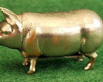 Könnte beinhalten: Kleine, metallische Schweinefigur mit eingerolltem Schwanz und roten Augen. Das Schwein steht auf vier Beinen, die Schnauze zeigt nach vorne. Die Figur hat eine glänzende, reflektierende Oberfläche und steht vor grünem Hintergrund.