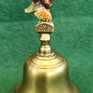 May include: A brass bell with a seated pixie figure on top. The pixie is brown and gold with green and orange accents. The bell has the words "Devon Pixies" on it.