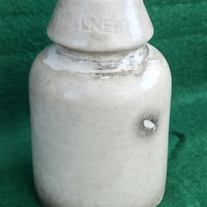 Op de afbeelding: Een crèmekleurige keramische fles met een afgeronde body en een conische top. De top is gemarkeerd met de letters "LNER". De fles vertoont tekenen van ouderdom, met wat verkleuring en een kleine donkere vlek. Vintage item.