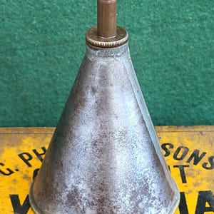 May include: A weathered metal oil can with a conical body and a brass spout. The can shows signs of age and use. The background features a yellow sign with the text "PHILLIPS HAND-CUT". The can is likely from the early 20th century.