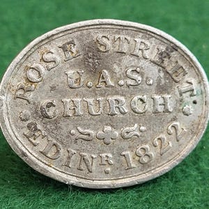 May include: An oval, silver-toned token with embossed text. It reads "ROSE STREET U.A.S. CHURCH EDIN 1822". There are decorative floral motifs. The token is displayed on a green surface.