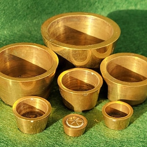 May include: Seven brass measuring cups of different sizes are displayed on a green surface. The cups have a shiny, golden finish and a simple, cylindrical design. The smallest cup features a star-like pattern. The cups are measured in metric units.