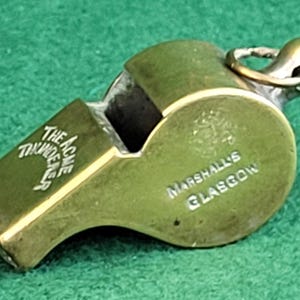 May include: A vintage brass whistle with the text "The Acme Thunderer" and "Marshalls Glasgow" engraved on its surface. The whistle has a loop for attaching a lanyard and is set against a green background.