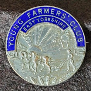 Puede incluir: Una insignia vintage plateada del Young Farmers' Club. El borde de esmalte azul dice "Young Farmers' Club East Yorkshire". El centro representa a un granjero arando con caballos, un sol y árboles.