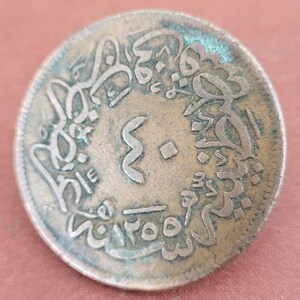 May include: A copper coin with Arabic script around the edge and a symbol in the centre. The coin is slightly tarnished and has a greenish patina.