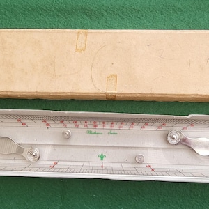 May include: Vintage slide rule with a clear plastic body and a beige cardboard box. The slide rule has red and black markings, with a silver-colored metal piece. The box is rectangular and has a worn appearance.