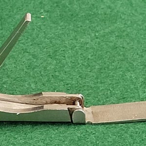 Może przedstawiać: Srebrny obcinacz do paznokci z dołączonym pilnikiem do paznokci. Obcinacz jest otwarty, z podniesioną dźwignią. Pilnik do paznokci jest wysunięty. Obcinacz do paznokci leży na zielonej powierzchni.