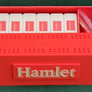 Puede incluir: Una caja de juego de plástico roja con una etiqueta blanca que dice "Hamlet" en la parte delantera. La caja tiene una cuadrícula de agujeros en la parte superior y un compartimento para las piezas del juego.