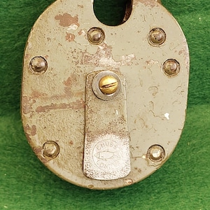 Peut inclure: Un cadenas ovale ancien et sa clé. Le cadenas est gris clair avec des signes d'usure et de rouille. La clé est en laiton. Le cadenas porte le mot "CHUBB" et un logo de poisson. Objet de sécurité vintage.
