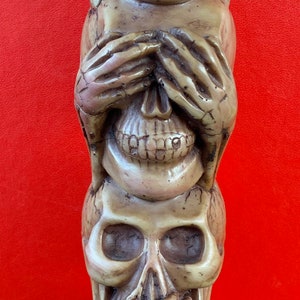 May include: A three-tiered sculpture of skulls, each with a different hand gesture representing the three wise monkeys: "See no evil, hear no evil, speak no evil". The bottom skull is holding its mouth closed, the middle skull is covering its eyes, and the top skull is covering its ears. The sculpture is made of a light brown material and has a rough, textured surface. The base of the sculpture is inscribed with the words "Do No Evil".