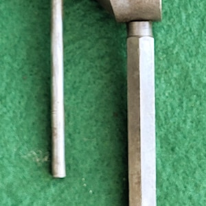 Peut inclure: Un outil en métal argenté avec une poignée en forme de T et un corps hexagonal. L'outil comporte une section filetée et une tête en forme de U. Le fond est de couleur verte unie.