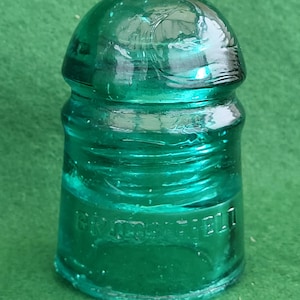 Pode incluir: Um isolador de vidro vintage, de cor verde-azulado, com um topo arredondado e uma base cilíndrica. O vidro tem um ligeiro padrão de redemoinho e a palavra "BROOKFIELD" está em relevo na lateral. Uma peça clássica de hardware elétrico antigo.