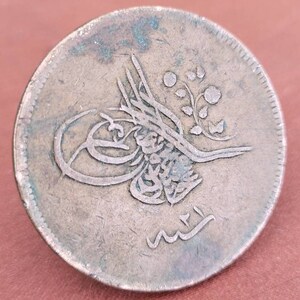 May include: A silver Ottoman coin featuring Arabic script and a floral design. The coin shows signs of wear and has a greenish patina.
