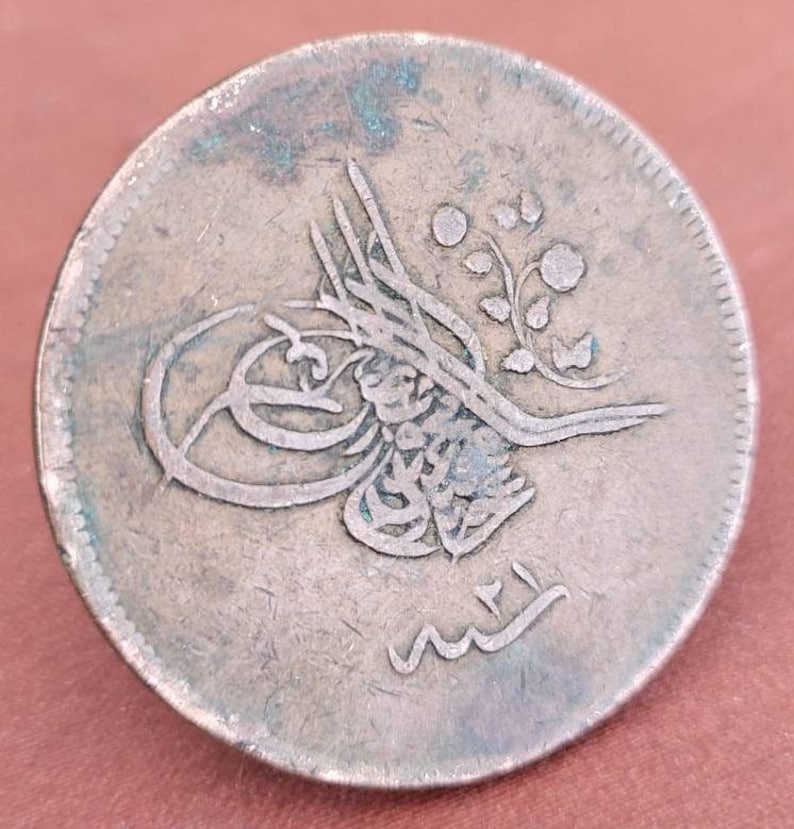 May include: A silver Ottoman coin featuring Arabic script and a floral design. The coin shows signs of wear and has a greenish patina.