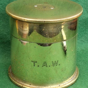 May include: A cylindrical brass container with a lid. The container has a polished, reflective surface and a slightly tarnished appearance. The initials "T.A.W." are engraved on the side. The lid has a circular indentation.
