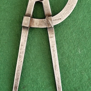 May include: Vintage metal compass with a curved arm and pointed tips. The compass is silver-toned and shows signs of age. The tool is designed for measuring and drawing circles. The background is a solid green.