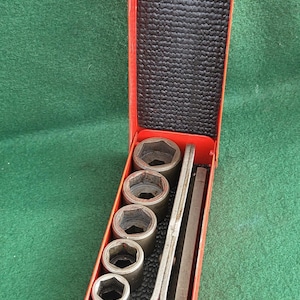 Puede incluir: Una caja de herramientas de metal roja, abierta para revelar un juego de llaves de vaso y una llave. Las llaves de vaso son de varios tamaños y la llave es plateada. El interior es negro y texturizado.