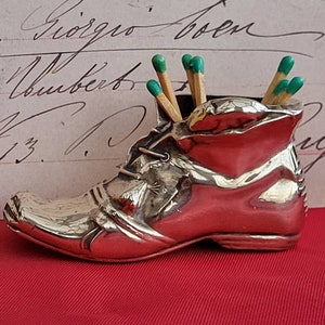 Puede incluir: Un portafósforos plateado en forma de bota con cerillas de punta verde en su interior. La bota tiene cordones y está sobre una superficie roja. El fondo presenta escritura manuscrita.
