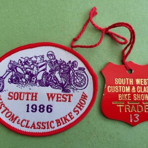 May include: A red and white oval patch with the text "South West Custom & Classic Bike Show 1986" and a cartoon image of three people riding motorcycles. A red shield-shaped tag with the text "South West Custom & Classic Bike Show Trade 13" is attached to the patch with a red string.