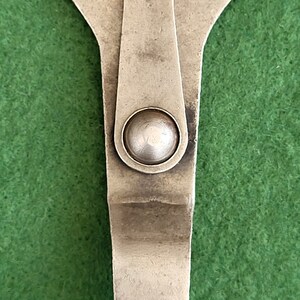 May include: A silver-coloured wrench with a rounded head and a small, round knob in the middle. The wrench has two prongs on the top and a square-shaped base.