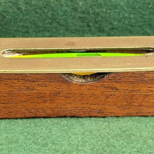 Puede incluir: Un nivel de latón con una base de madera. El nivel tiene una burbuja verde y está marcado con las palabras "Made in England".