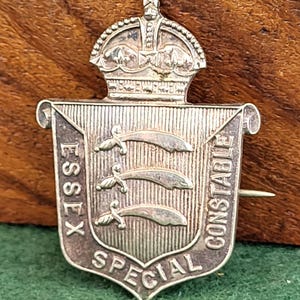 Puede incluir: Una insignia plateada de Constable Especial de Essex. La insignia presenta una corona en la parte superior, un escudo con tres espadas estilizadas y las palabras "Essex Special Constable" alrededor de los bordes. La insignia tiene un alfiler en la parte posterior.