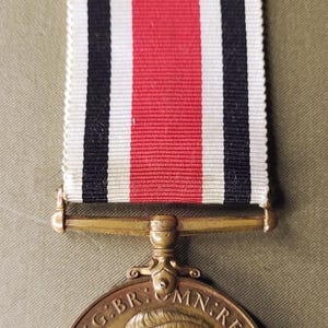 Puede incluir: Medalla vintage con acabado bronce, con perfil de hombre y texto. La cinta tiene rayas verticales negras, blancas y rojas. La medalla es probablemente un artículo conmemorativo.