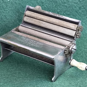 May include: A vintage metal cigarette rolling machine with a hand crank and a white handle. The device features three grooved metal rollers and is engraved with the text THE LITTLE DON SOLE MAKER EVANS PATENTEE LONDON on the top surface.