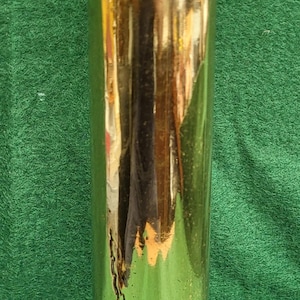 May include: A tall, cylindrical brass object with a shiny, reflective surface. The object has a rounded base and a slightly wider top. It stands against a green background.