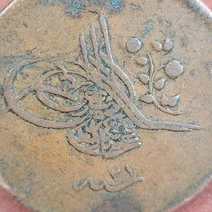 May include: Close-up of a copper coin featuring Arabic script and a floral design. The coin is worn and has a green patina.