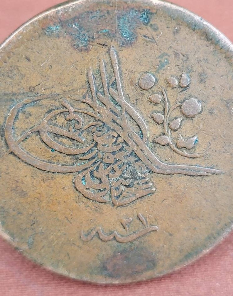 May include: Close-up of a copper coin featuring Arabic script and a floral design. The coin is worn and has a green patina.