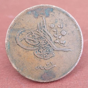 May include: A copper coin featuring Arabic script and a floral design. The coin is worn and has a green patina.