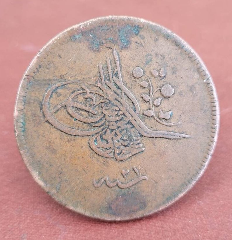 May include: A copper coin featuring Arabic script and a floral design. The coin is worn and has a green patina.