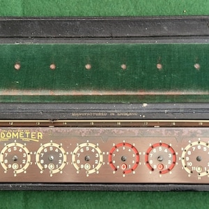 Peut inclure: Un calculateur Addometer vintage ouvert dans un étui en cuir noir. Le calculateur comporte plusieurs cadrans numérotés avec des accents rouges. L'étui a un intérieur en feutre vert et est ouvert sur une surface verte.