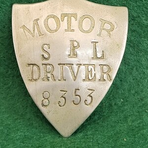 May include: A vintage silver-coloured shield-shaped badge. The badge has the words "MOTOR SPL DRIVER" and the number "8353" embossed on it. The badge is set against a green background.