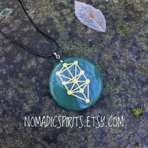 May include: A green round pendant with a gold geometric design, hanging from a black cord. The pendant is on a gray textured surface. NOMADICSPIRITS.ETSY.COM is written below the pendant.