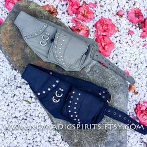 以下が含まれることがあります： 調節可能なストラップ付きのファブリックヒップバッグ2つ。1つはダークブルー、もう1つはグレー。両方のバッグには複数のポケットがあり、シルバーのスタッズとメタルリングで飾られています。