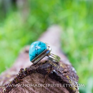 May include: A turquoise gemstone ring set in silver. The ring is on a wooden surface with a green background.