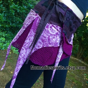 May include: A purple and white patterned skirt with a brown fabric belt tied around the waist. The skirt has a crocheted layer and a fringe detail. The text "Nomadcspirits.etsy.com" is visible in the image.