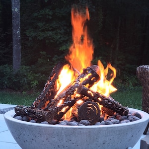 May include: A concrete fire pit with a large, orange fire burning inside. The fire pit is round and has a textured surface. The fire is surrounded by small, black stones.
