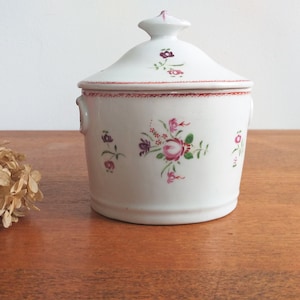Puede incluir: Azucarero de cerámica blanca con tapa, decorado con diseños florales rosas y verdes. El tazón tiene dos pequeñas asas a los lados.