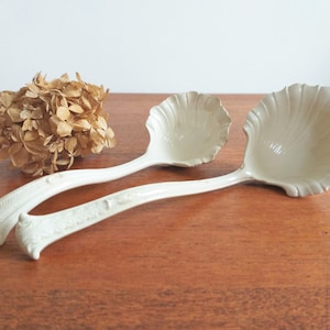 May include: Two white ceramic soup ladles with a scalloped design. The ladles have ornate handles and are on a wooden surface.