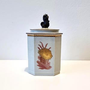 May include: A pale blue, hexagonal ceramic jar with a lid and a black finial. The jar features a gold and red floral design. The lid has a gold trim. The jar is on a white surface.
