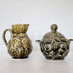 May include: A green ceramic creamer and sugar bowl set with a floral design. The creamer has a handle and a spout. The sugar bowl has a lid and a knob on top.