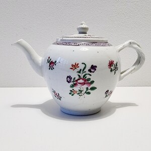 Puede incluir: Tetera de porcelana blanca con diseño floral. La tetera presenta un pico curvo, un asa retorcida y una tapa con un pequeño pomo. El patrón floral incluye flores rosas, moradas y verdes. Un patrón geométrico morado y blanco decora el borde de la tapa.