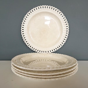 May include: A set of off-white ceramic plates with a scalloped, openwork border. One plate is displayed above a stack of four similar plates. The plates have a slightly textured surface and a glossy finish, suitable for serving food.