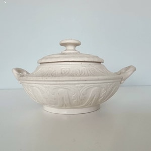 Può includere: Un piatto da portata in ceramica bianca con coperchio e due manici. Il piatto è decorato con un motivo floreale in rilievo.