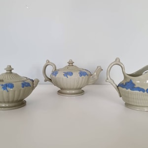 Puede incluir: Un juego de té vintage con tetera, jarra de crema y azucarero. El juego está hecho de cerámica blanca con diseños florales azules.
