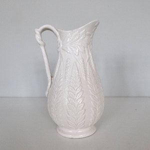 Può includere: Brocca in ceramica bianca con un motivo a felce. La brocca ha un manico con un design attorcigliato.